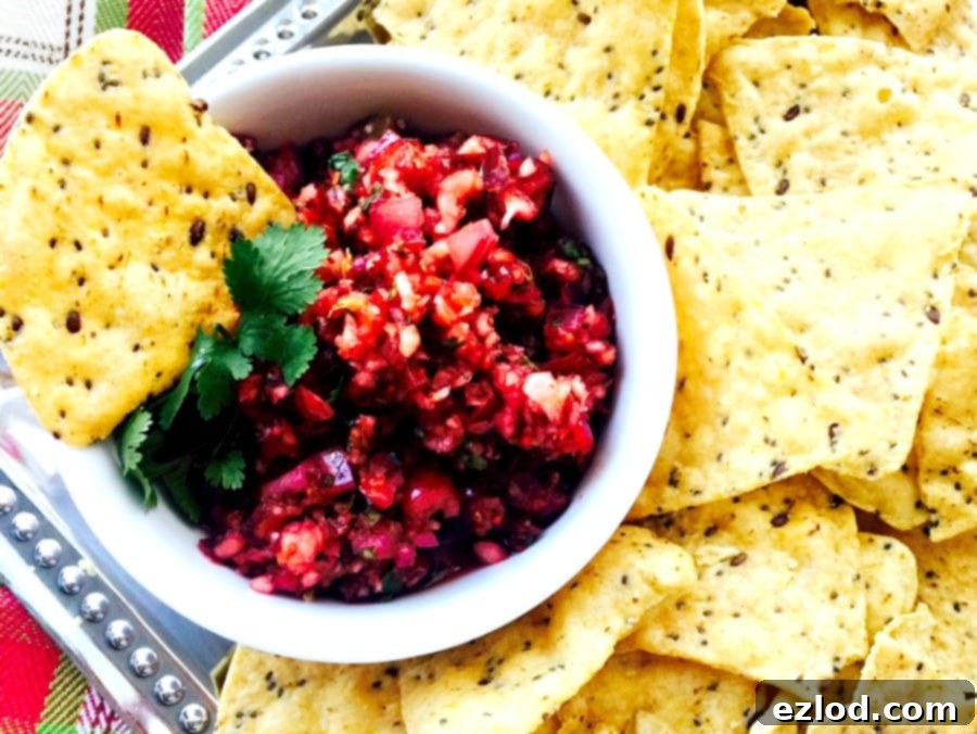 Close up photo of Holiday Salsa in a white bowl with tortilla chips on a silver platter. A festive green and red towel is in the background.