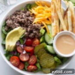 Burger bowl with ground beef, tomatoes, pickles, lettuce, sliced avocado, fries, and a side of special sauce.