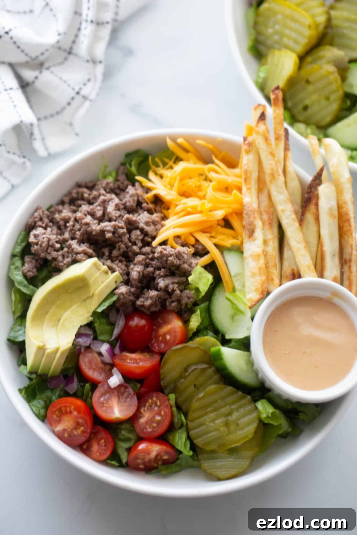 Burger bowl with ground beef, tomatoes, pickles, lettuce, sliced avocado, fries, and a side of special sauce.