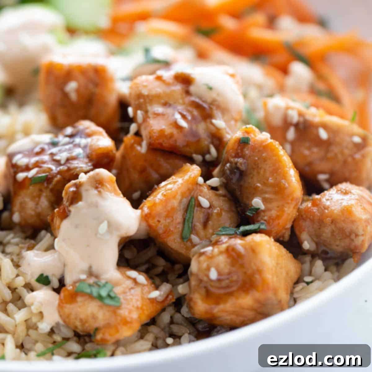 A vibrant bowl featuring golden-brown sticky air fryer salmon bites, nestled over a bed of fluffy brown rice, generously drizzled with spicy mayo, and garnished with toasted sesame seeds and fresh cilantro.