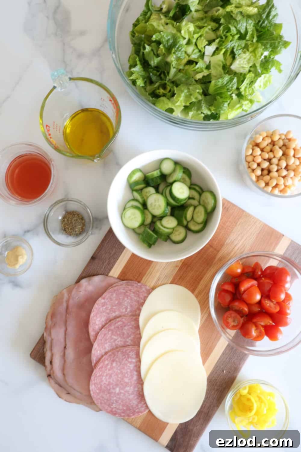 TikTok's Favorite Italian Grinder Chopped Salad 3 Individual fresh ingredients laid out on a white countertop, ready to be assembled into an Italian Chopped Grinder Salad, including various deli meats, lettuce, tomatoes, cucumbers, and pepperoncini.