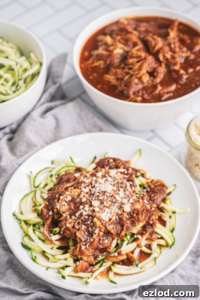 A white plate of balsamic chicken over spiralized zoodles and topped with parmesan cheese. In the background are two large bowls, one is full of plain zoodles, the other is full of balsamic chicken and sauce.
