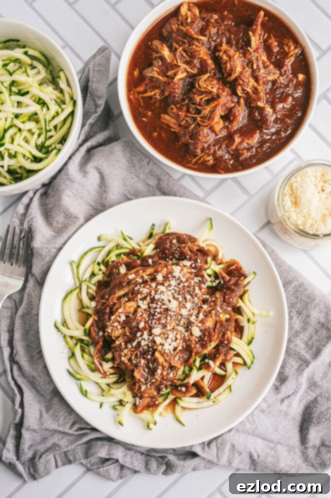 Overhead photo of balsamic chicken with zoodles on a white plate. At the top of the photo are two bowls, one filled with zoodles, the other filled with balsamic chicken. There is a small glass jar filled with parmesan cheese, too.