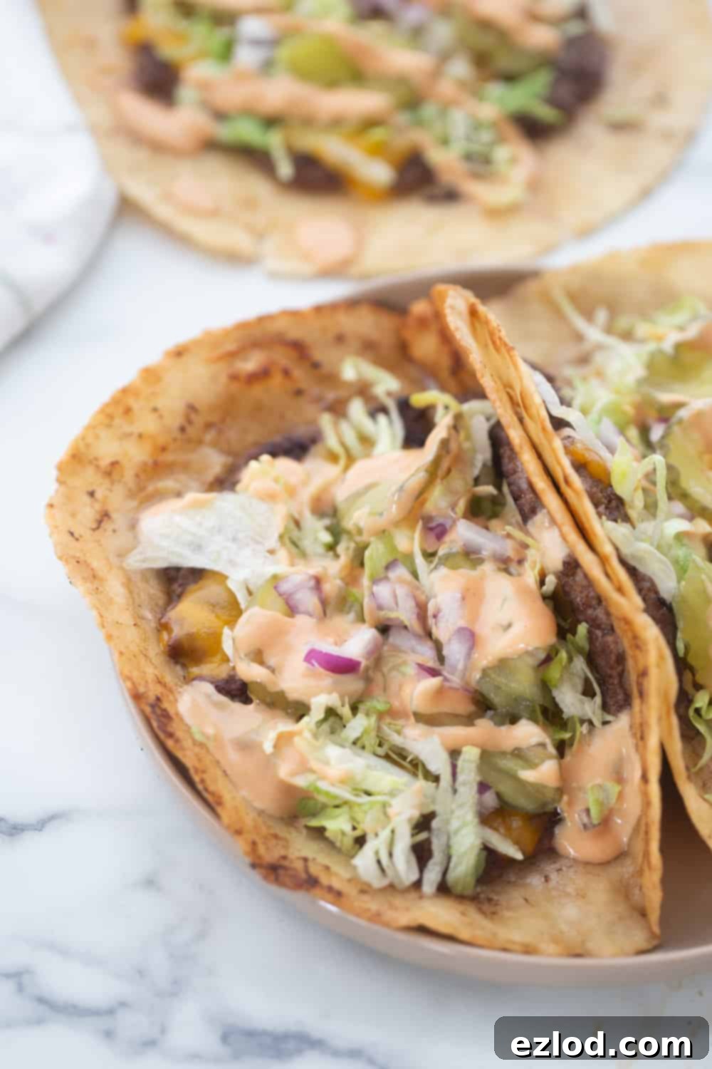 A stunning top-down view of two perfectly assembled gluten-free Big Mac tacos on a minimalist white plate, overflowing with fresh lettuce, creamy special sauce, vibrant pickles, and crisp onions.
