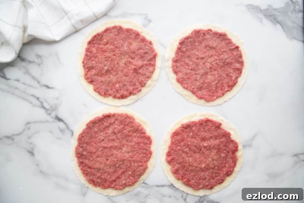 Four pristine gluten-free tortillas laid out, each meticulously topped with a thin, unseasoned raw ground beef patty, pressed almost to the very edge, awaiting its culinary transformation.