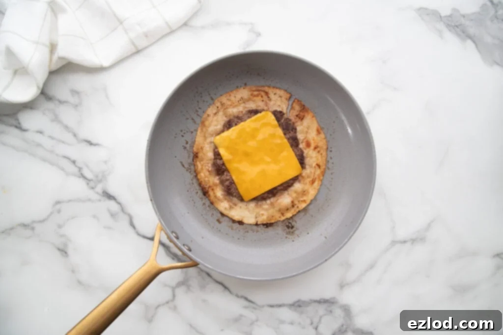 A gluten-free tortilla in a pan, expertly flipped with the beef patty facing up, now crowned with a perfectly melting slice of American cheese, gently steaming under a lid.