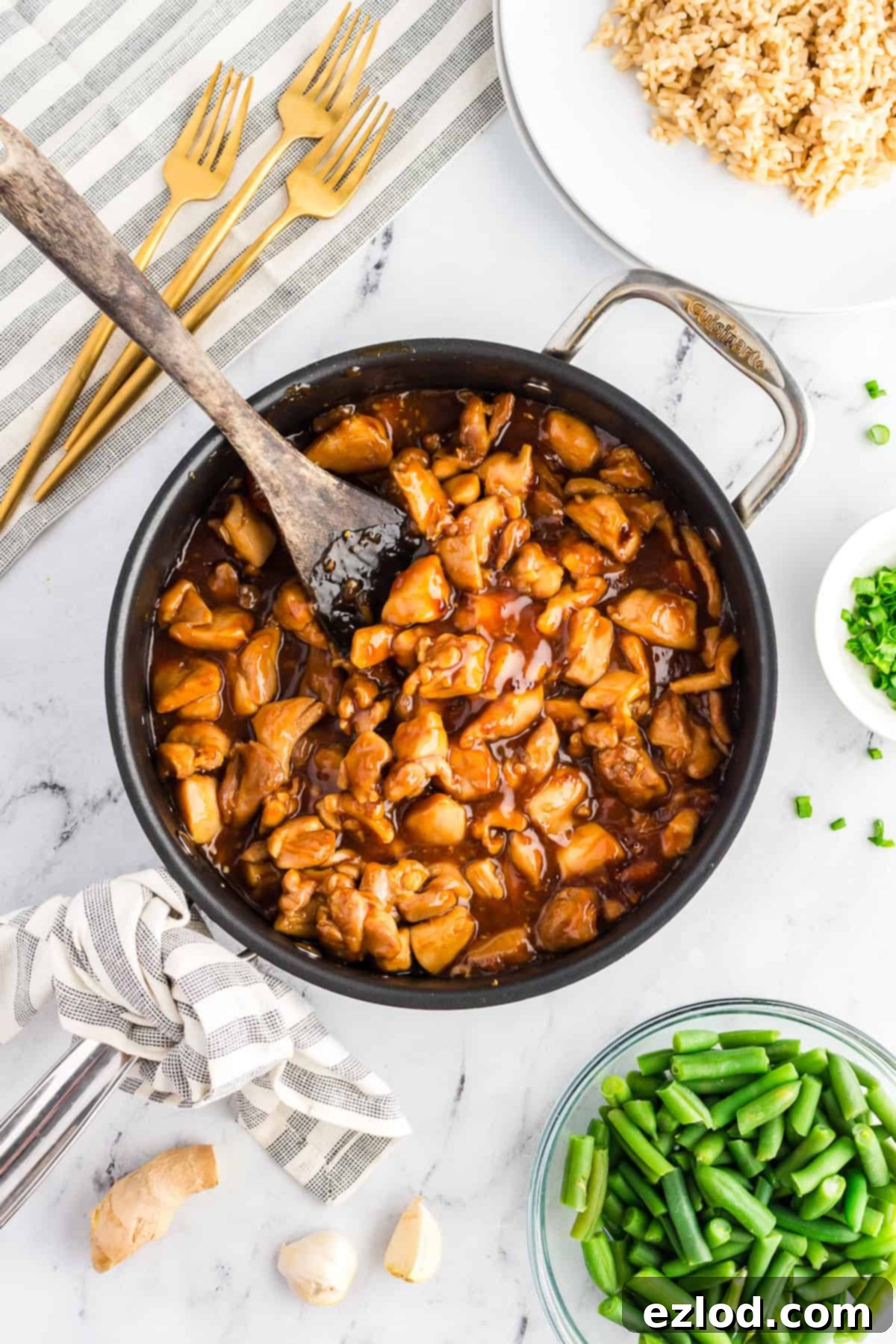 A cast iron skillet filled with homemade bourbon chicken, served alongside a bowl of brown rice, a side of green beans, and a small dish of chopped green onions for garnish.