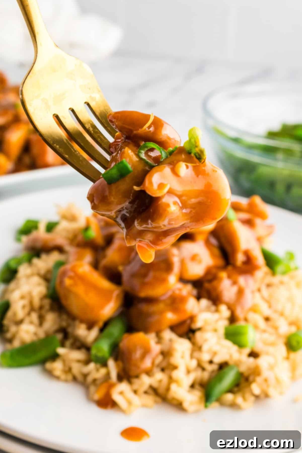A close-up of a fork taking a bite of bourbon chicken over brown rice, served with green beans and garnished with green onions.