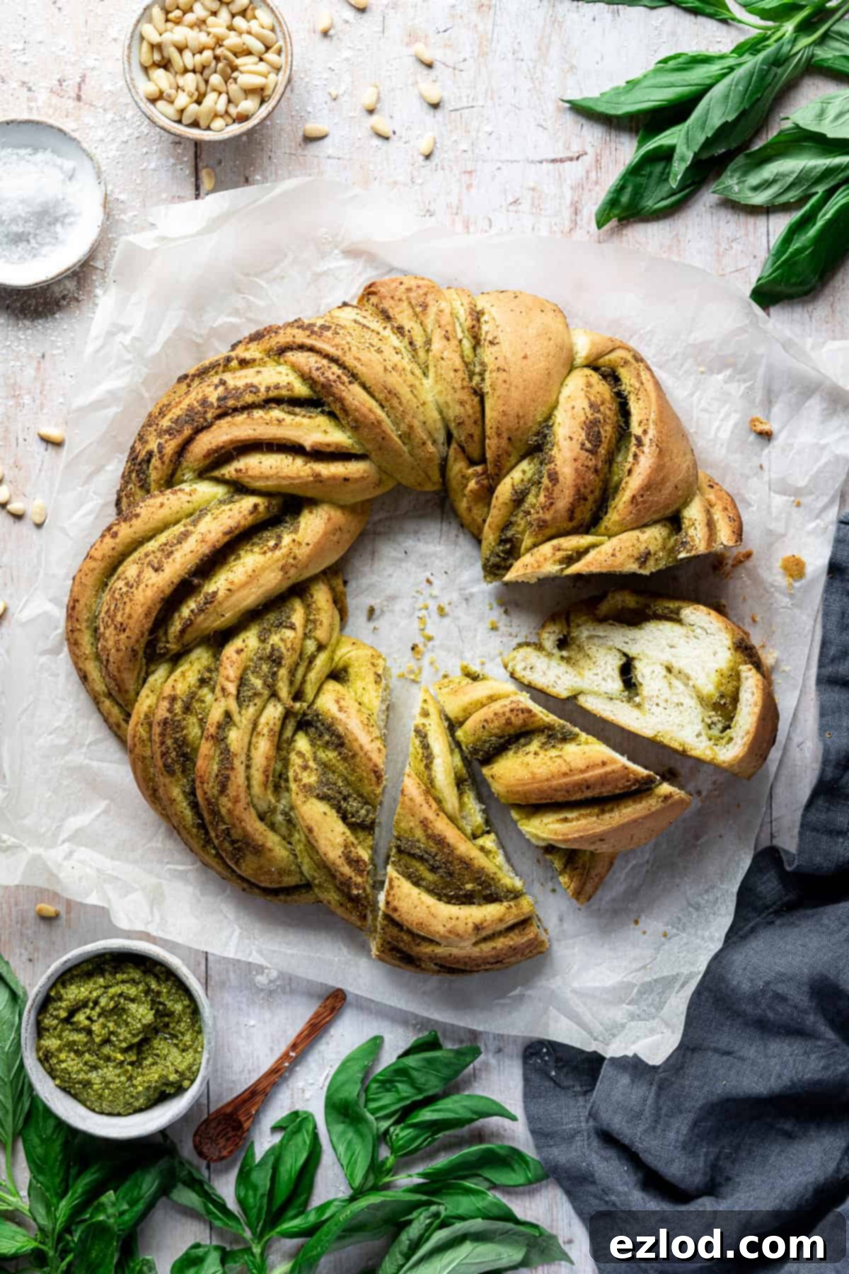 A beautifully braided vegan pesto bread wreath with three slices already cut, resting on a rustic light wooden table. It's elegantly surrounded by fresh green basil leaves, golden pine nuts, coarse sea salt, and a small bowl of vibrant green pesto, highlighting its key ingredients and fresh appeal.