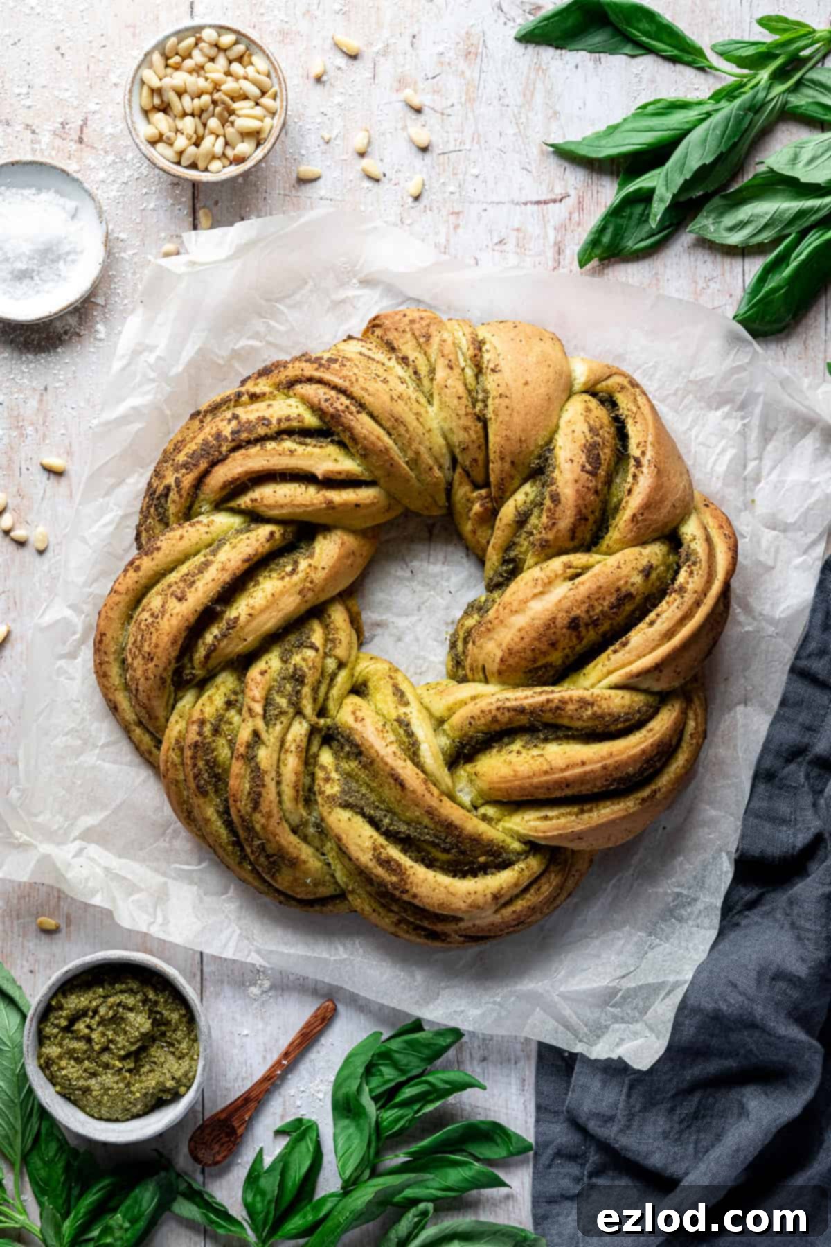 A freshly baked vegan pesto bread wreath, golden-brown and beautifully braided, resting on a sheet of baking parchment on a rustic wooden table. It is surrounded by fresh basil leaves, pine nuts, coarse salt, and a bowl of pesto, evoking a sense of homemade goodness and fresh ingredients.