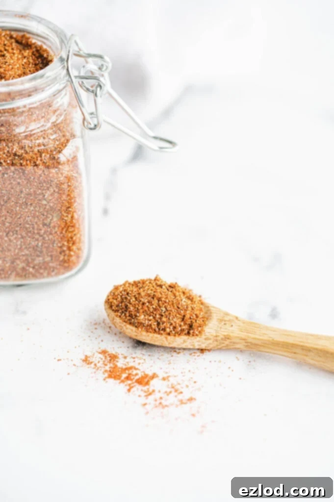 A glass jar filled with homemade taco seasoning blend, accompanied by a small wooden spoon overflowing with the rich, reddish-brown spice mix, all set on a bright white surface.