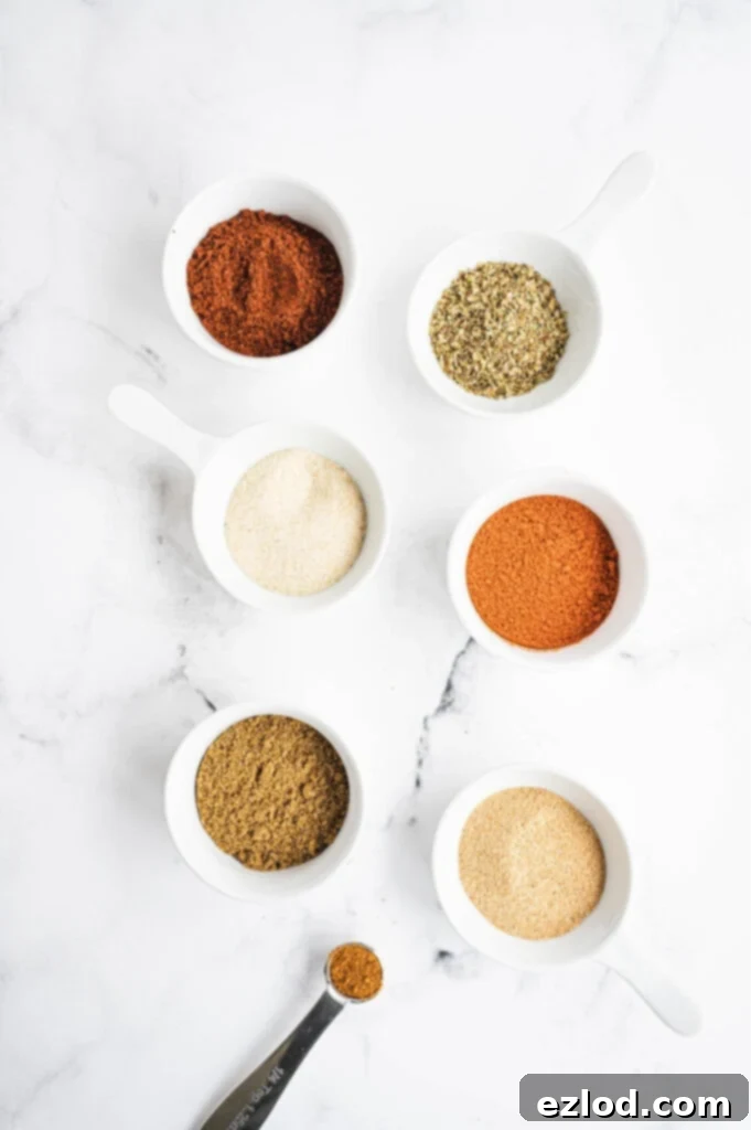 An overhead shot showcasing individual small white bowls filled with various vibrant spices like red chili powder, orange paprika, brown cumin, and white garlic powder, arranged neatly on a light surface with a measuring spoon, ready to be blended into homemade taco seasoning.