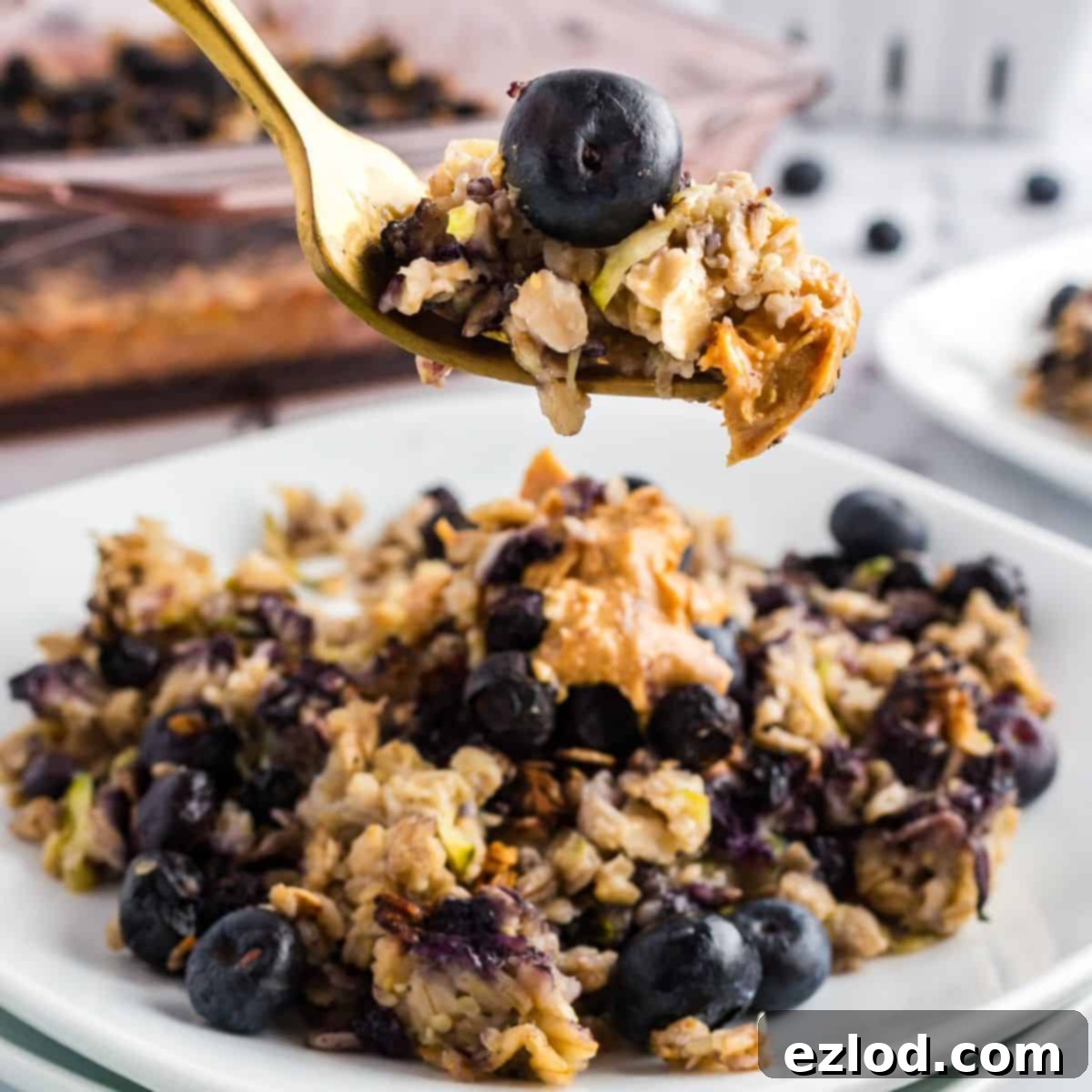 A serving of zucchini blueberry baked oatmeal on a white plate, garnished with fresh blueberries and a gold fork.