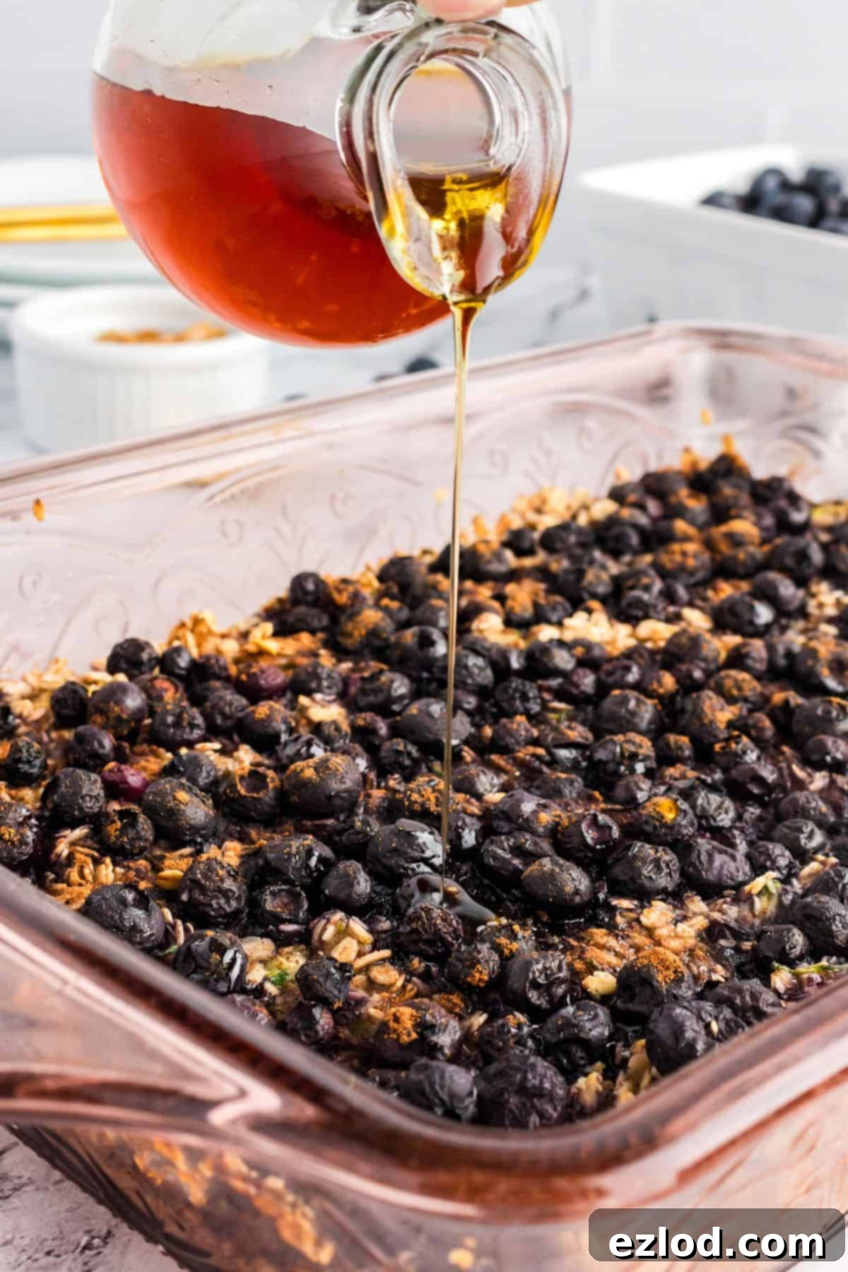 A glass jug pouring maple syrup over a freshly baked dish of zucchini blueberry oatmeal.