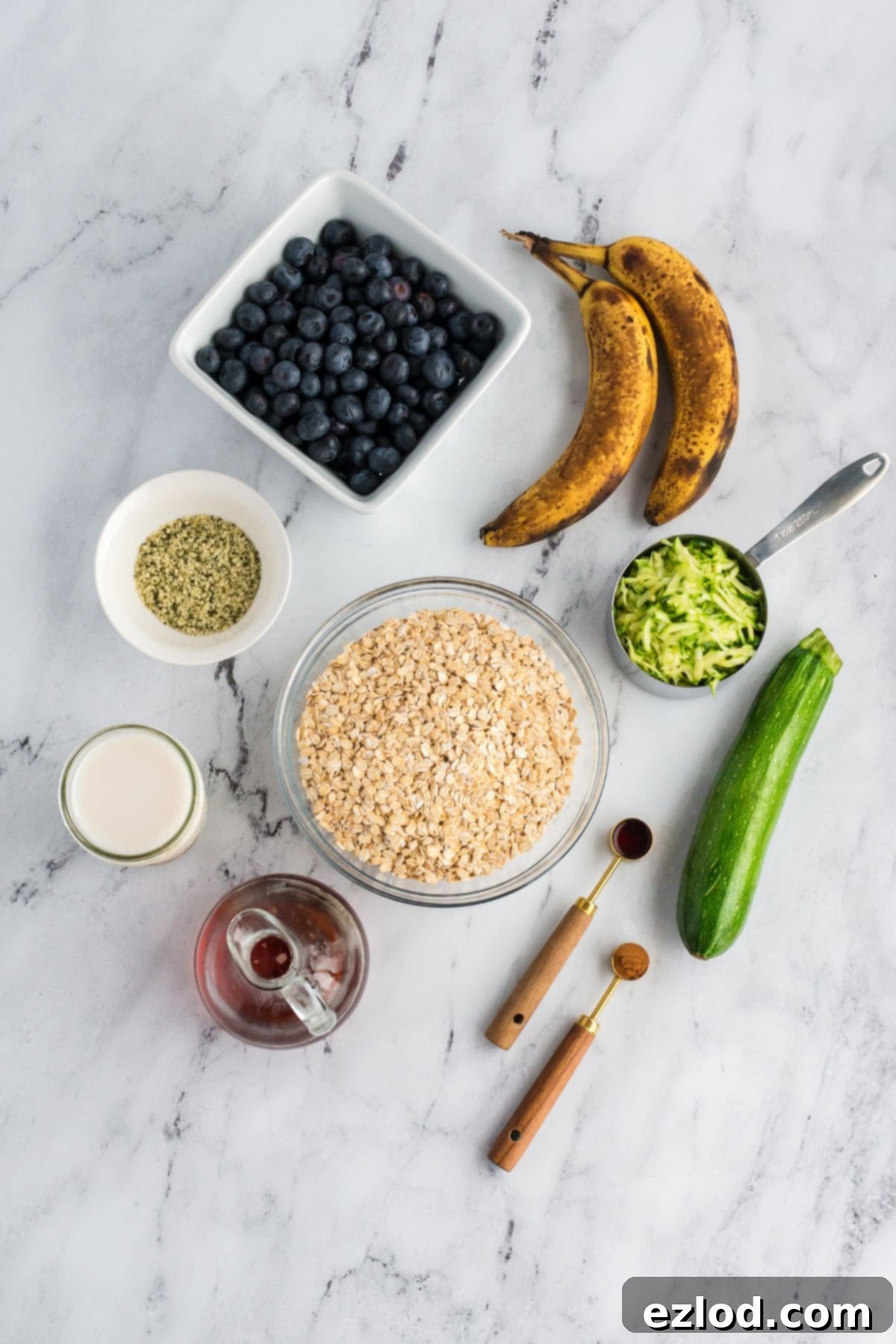 Ingredients for zucchini blueberry baked oatmeal including oats, blueberries, bananas, zucchini, milk, and spices arranged on a marble surface.