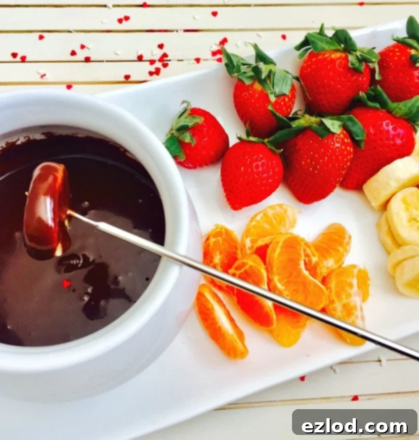 White fondue pot filled with chocolate with a mandarin orange segment being dipped in by a metal fondue fork. On the side of the rectangular serving dish are whole strawberries, sliced bananas, and mandarin orange segments. 