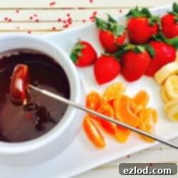 A small white pot of dairy free chocolate fondue on a white rectangular serving dish with whole strawberries, sliced bananas, and mandarin orange segments.