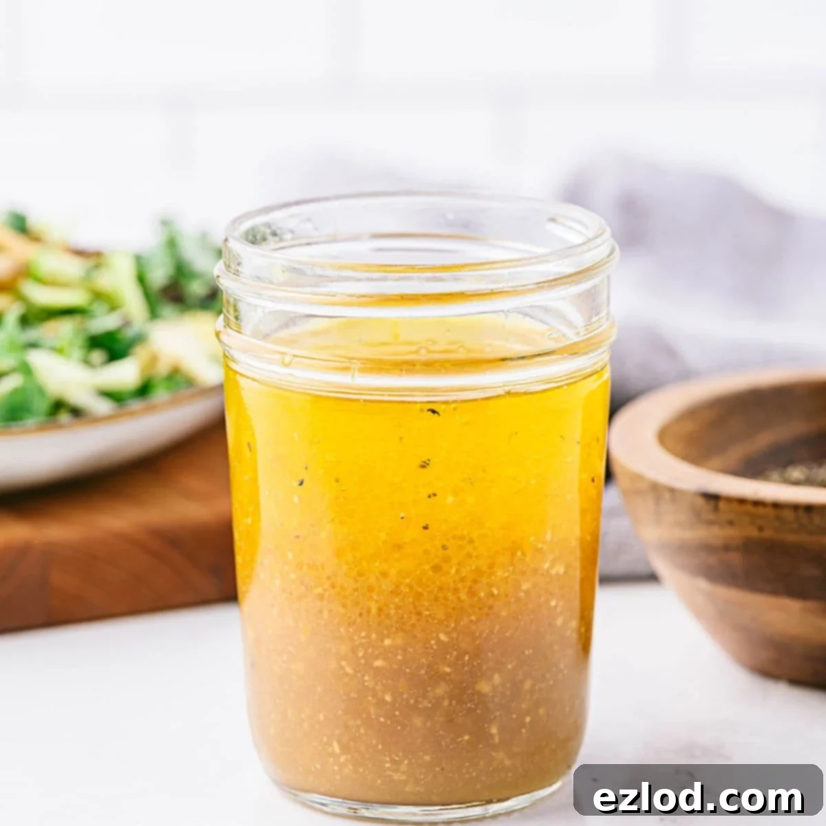 A jar of freshly mixed apple cider vinaigrette, with the layers of oil and vinegar starting to combine. A salad and wooden bowl with pepper are visible in the background.