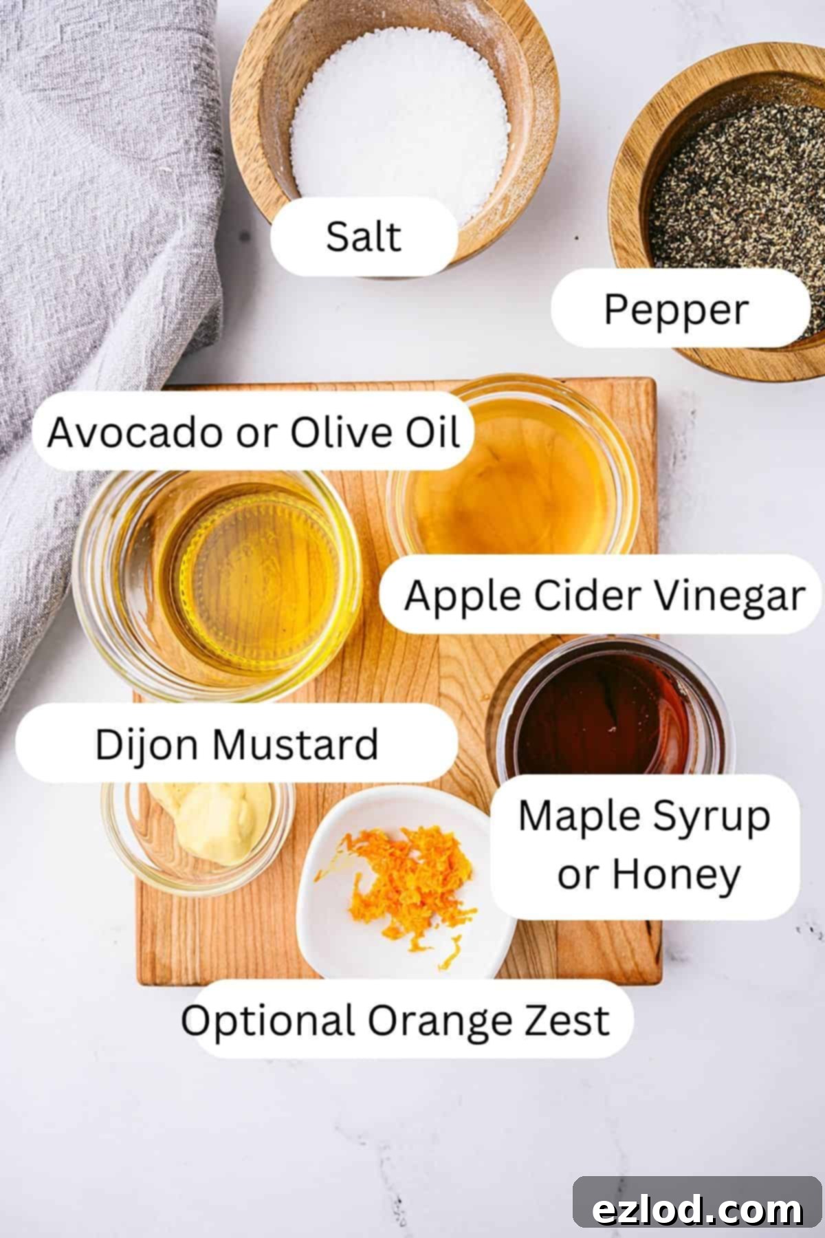 Ingredients for apple cider vinaigrette on a wooden board, including bowls of salt, pepper, avocado or olive oil, apple cider vinegar, Dijon mustard, maple syrup or honey, and optional orange zest.
