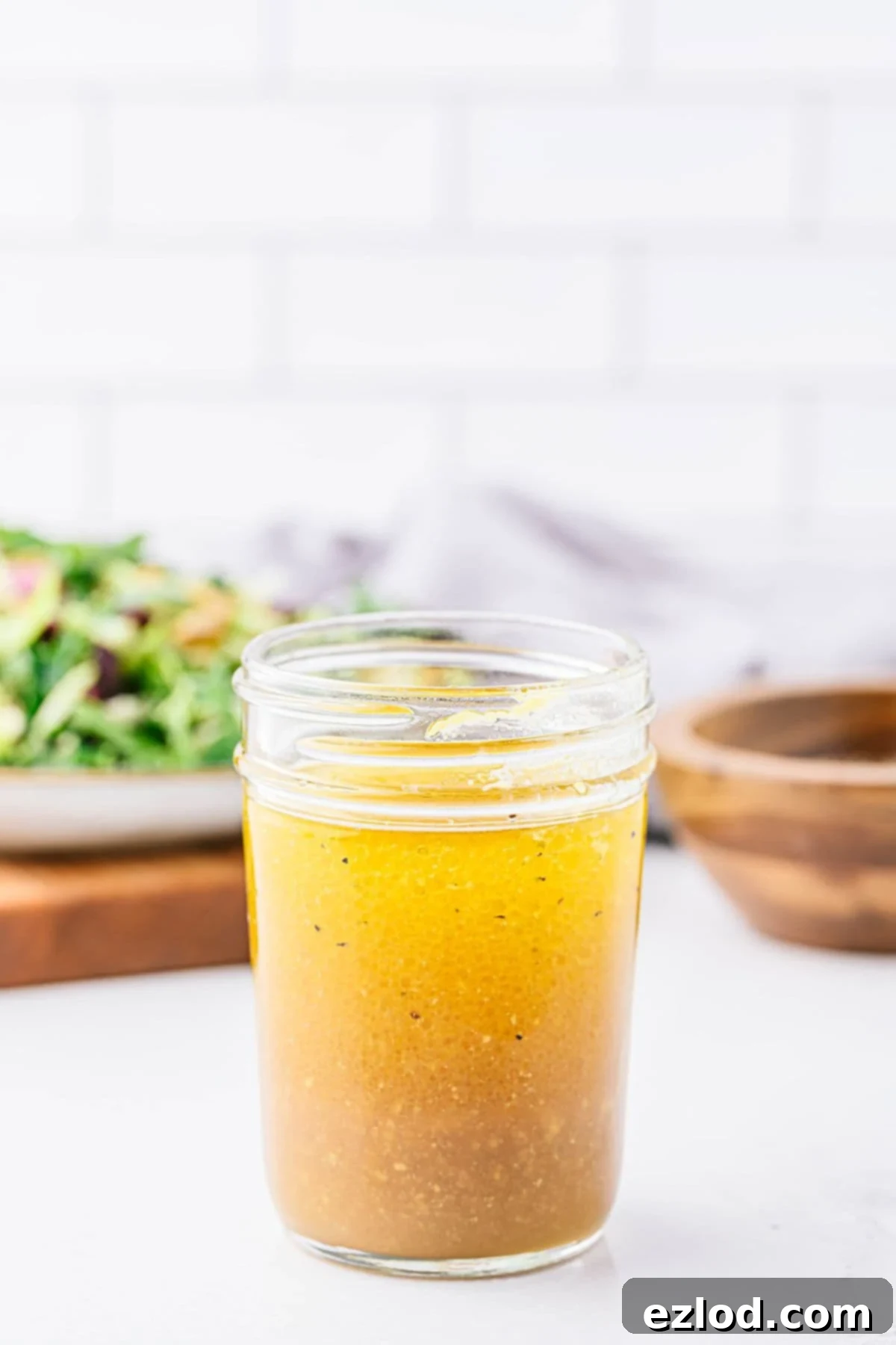 vinaigrette, with the layers of oil and vinegar starting to combine. A salad and wooden bowl with pepper are visible in the background.