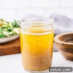 A jar of freshly mixed apple cider vinaigrette, with the layers of oil and vinegar starting to combine. A salad and wooden bowl with pepper are visible in the background.