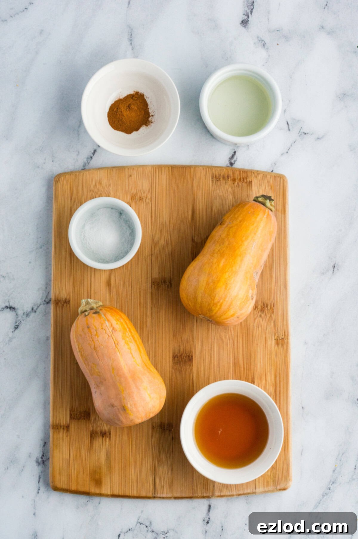 Sweet Maple Roasted Honeynut Squash Air Fryer Oven 4 A rustic wooden cutting board displays all the essential ingredients for roasted honeynut squash: whole honeynut squash, a small bowl of ground cinnamon, a sprinkle of sea salt, a bottle of olive oil, and a jar of pure maple syrup, ready for preparation.