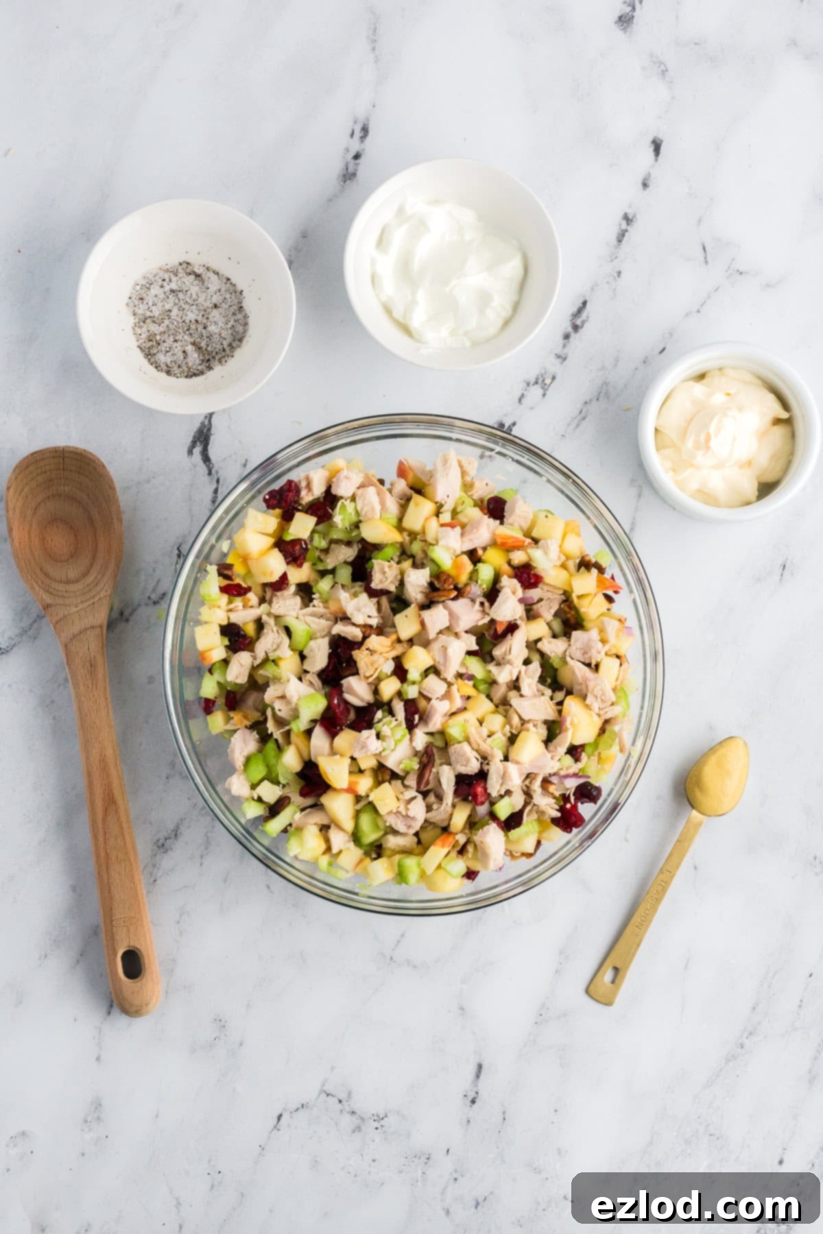 A large clear glass mixing bowl contains the chopped turkey meat, diced apples, celery, red onion, and pecans, ready for the dressing to be added, on a white marble table. Smaller bowls with mayonnaise, Greek yogurt, and seasoning are neatly arranged around it, with a wooden spoon nearby.