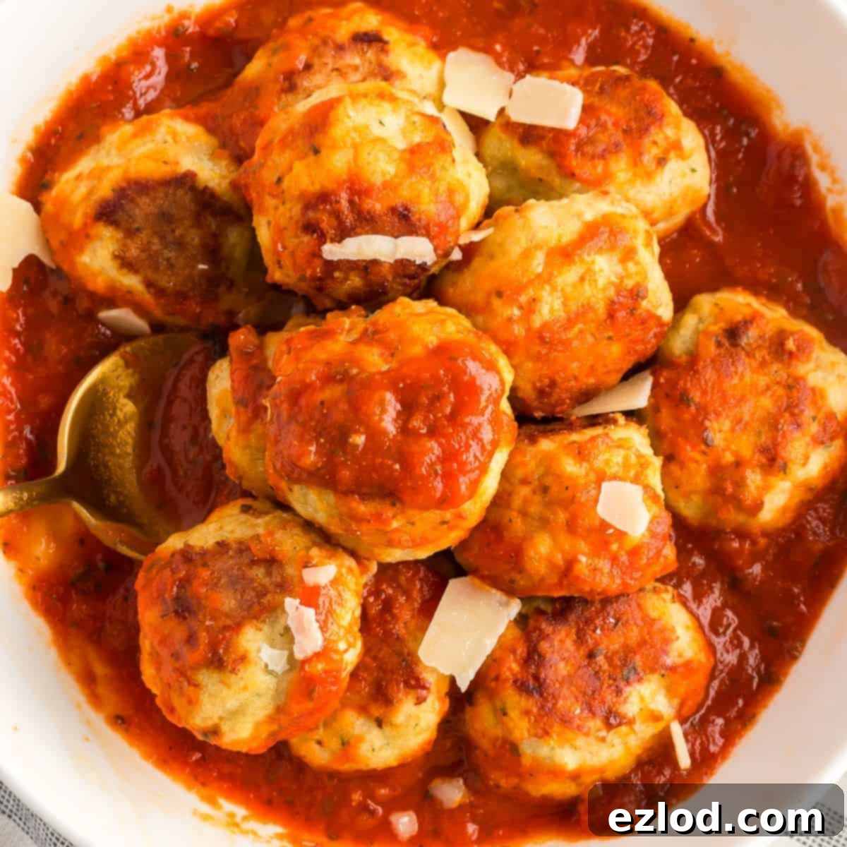 Baked chicken meatballs in a white bowl of sauce topped with shaved parmesan cheese.