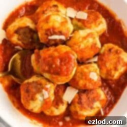Baked chicken meatballs in a white bowl of sauce topped with shaved parmesan cheese.