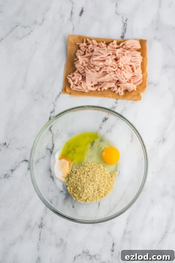 Glass bowl with eggs and breadcrumb sitting next to lean ground chicken on parchment paper.