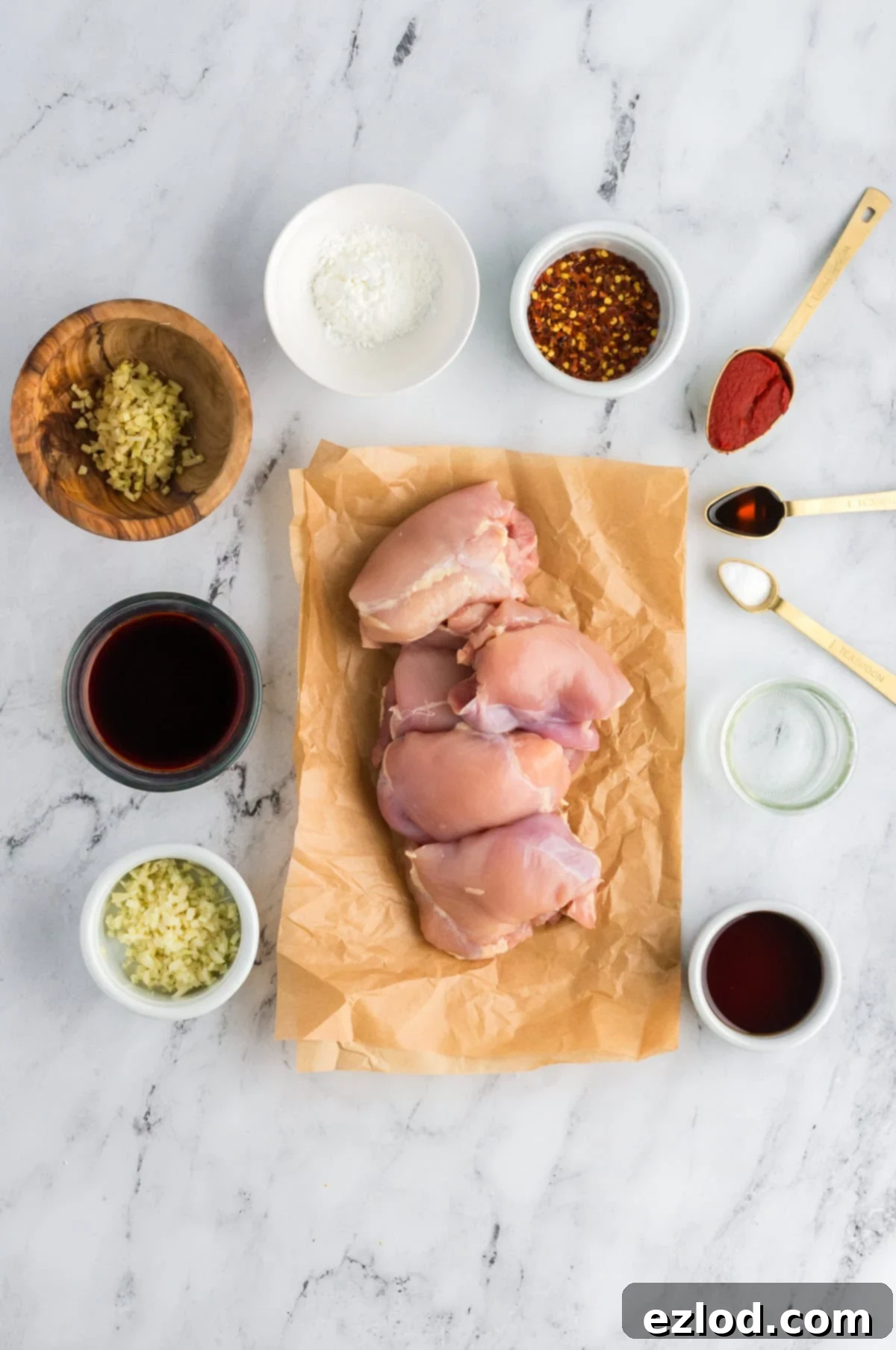 Glazed Sticky Chicken Thighs: Air Fried or Oven Baked 4 raw boneless skinless chicken thighs on parchment paper all on a marble background with all the ingredients for the marinade and sauce in separate containers around them.