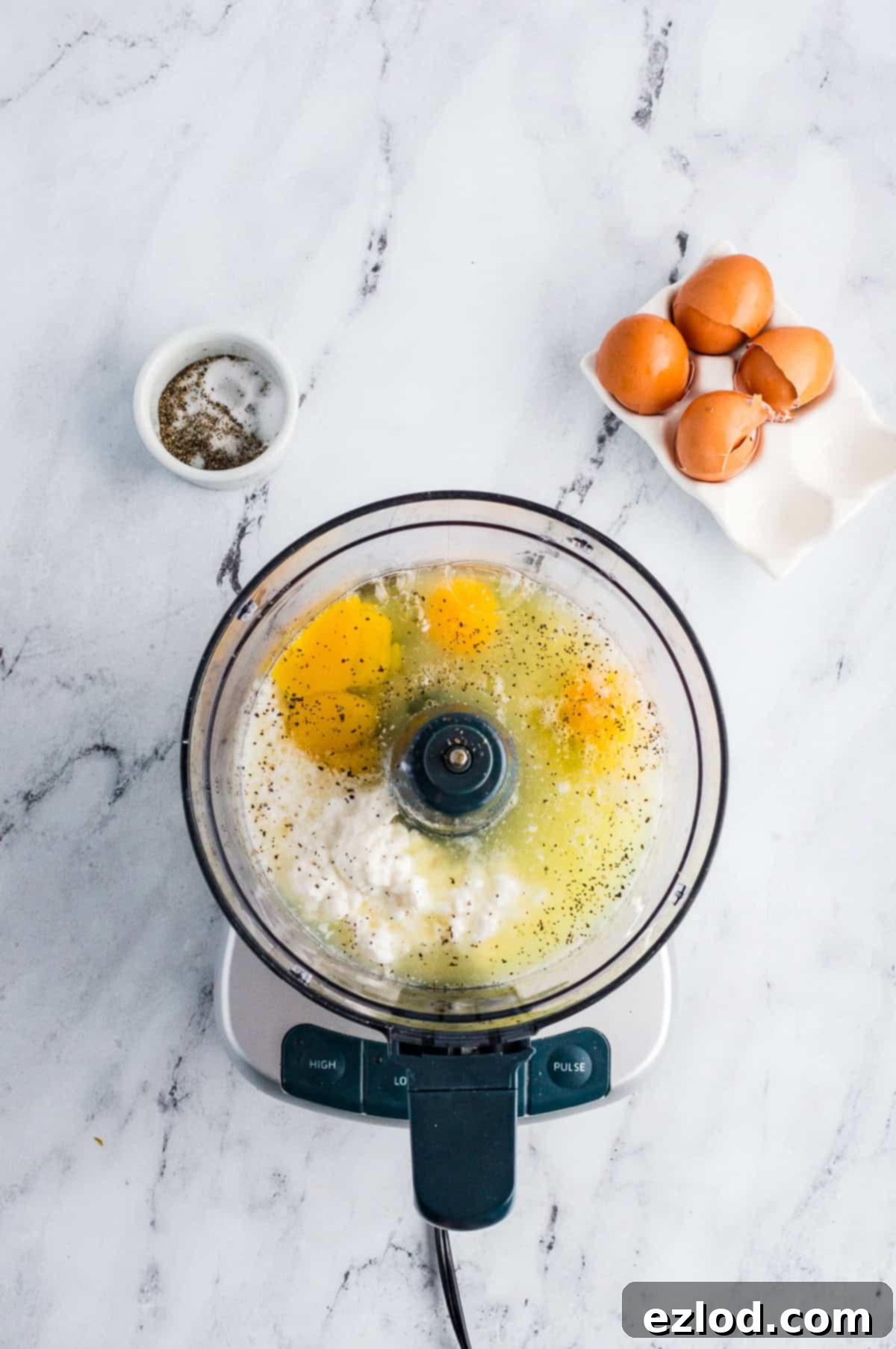 Eggs, cottage cheese, and salt and pepper in a food processor on a marble background before being blended.