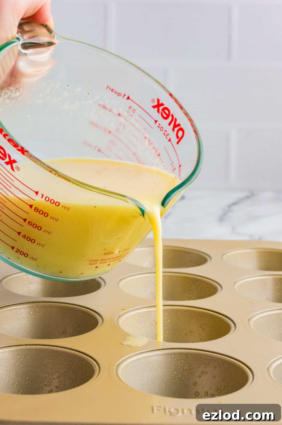 A glass measuring cup pouring the mixed egg bites mixture into the prepared muffin tins.