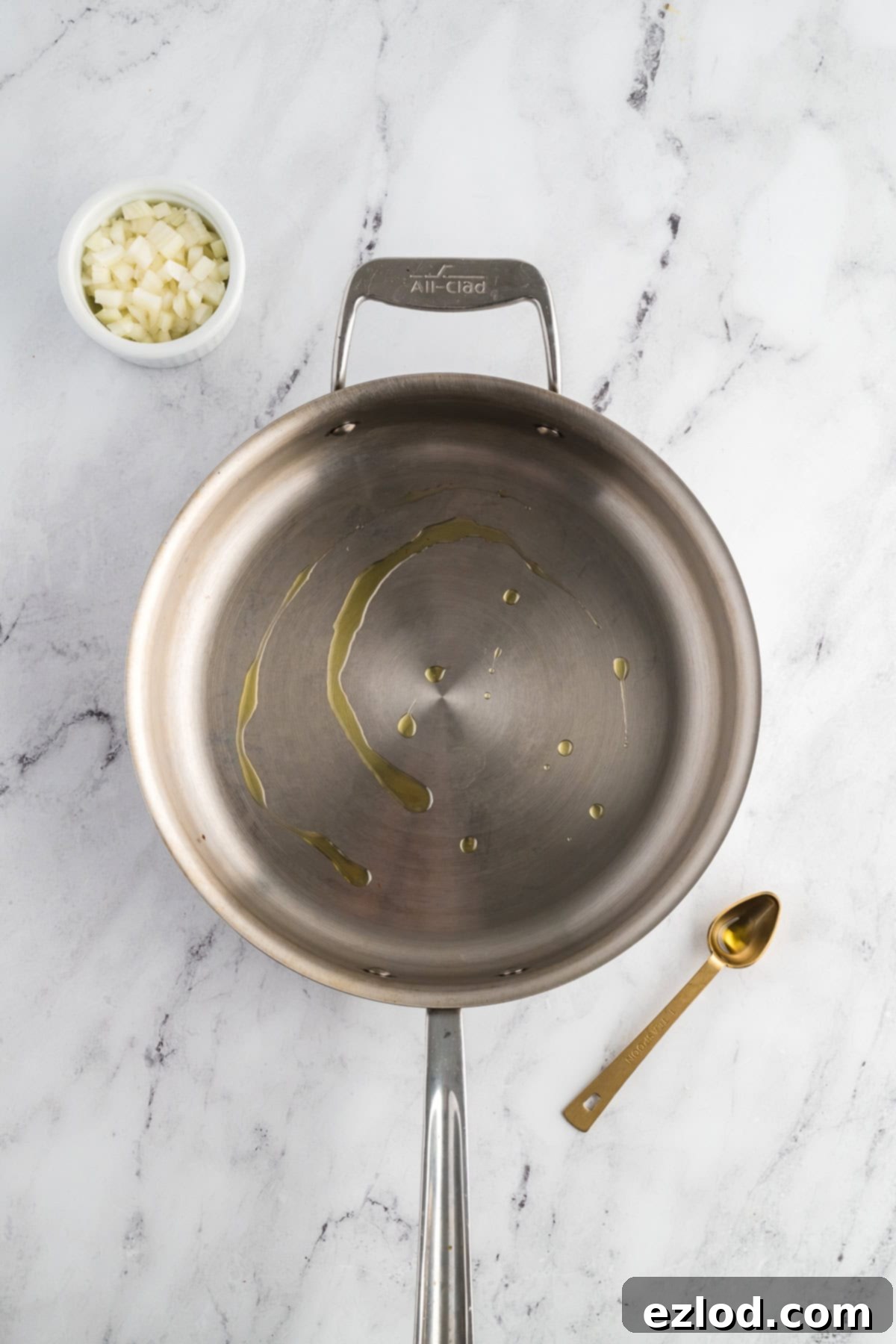 A skillet with olive oil shimmering over medium heat, with finely diced onions in a separate small bowl on a marble countertop.