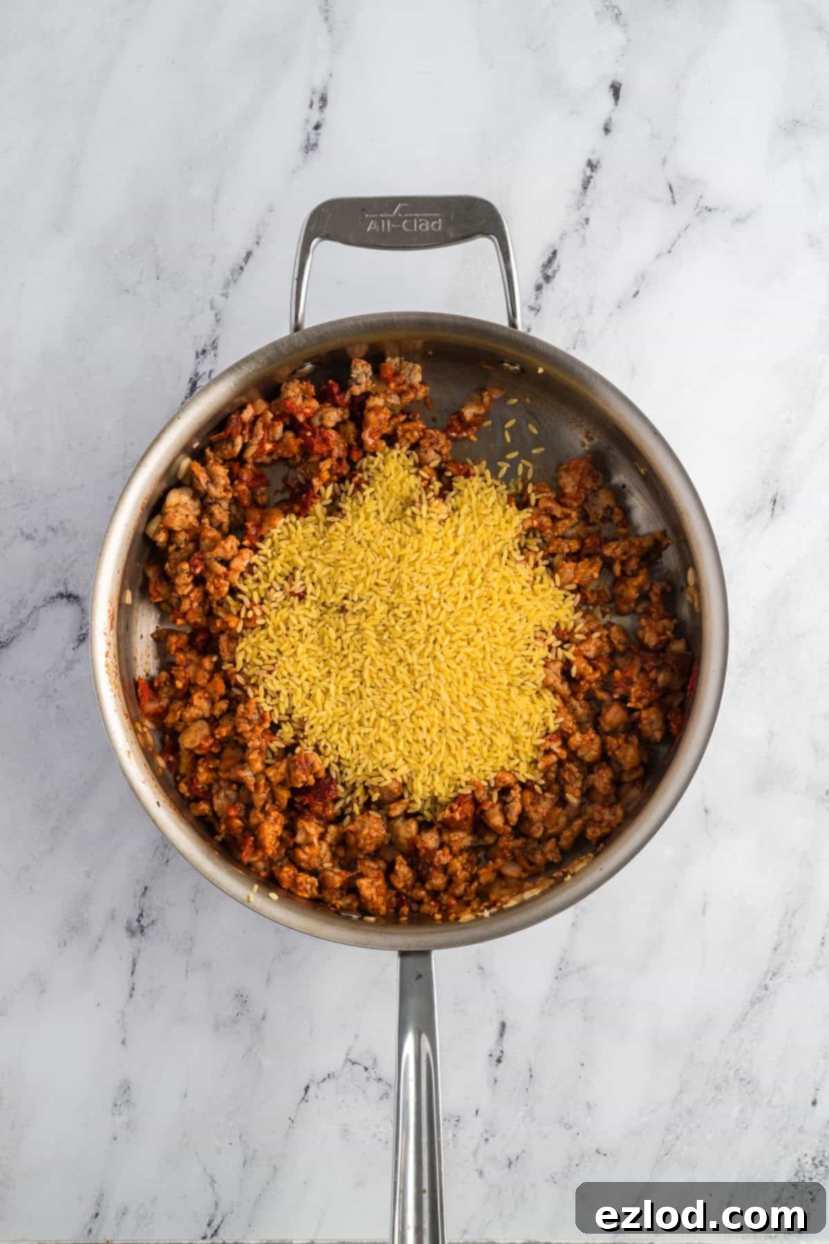 Gluten-free orzo pasta being added to the skillet containing cooked chicken sausage, onions, garlic, sun-dried tomatoes, and tomato paste.