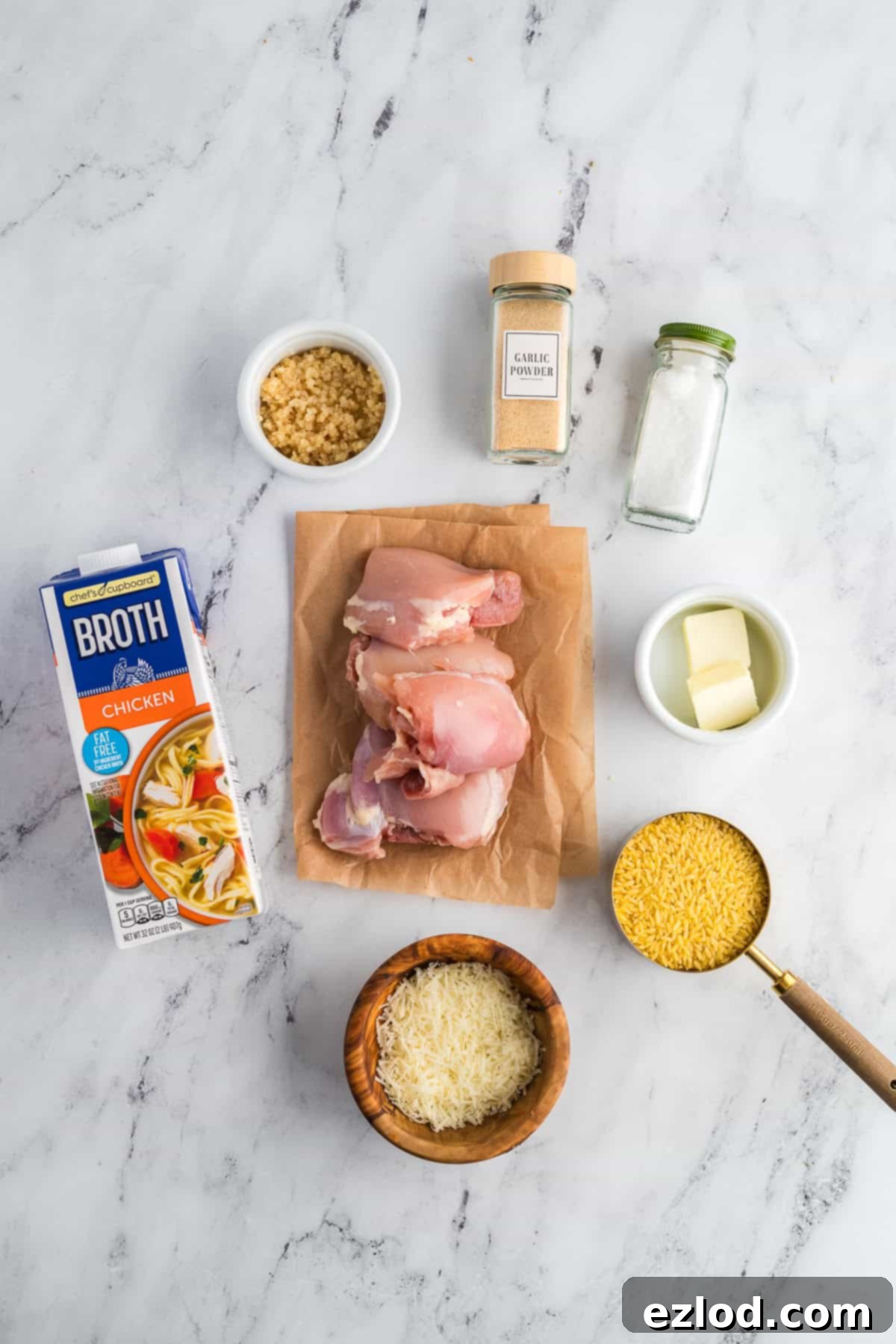 Your Go To One Pan Chicken Orzo 3 Ingredients for chicken and orzo skillet on a marble background in separate containers, including butter, garlic, chicken, orzo, broth, parmesan, and parsley.