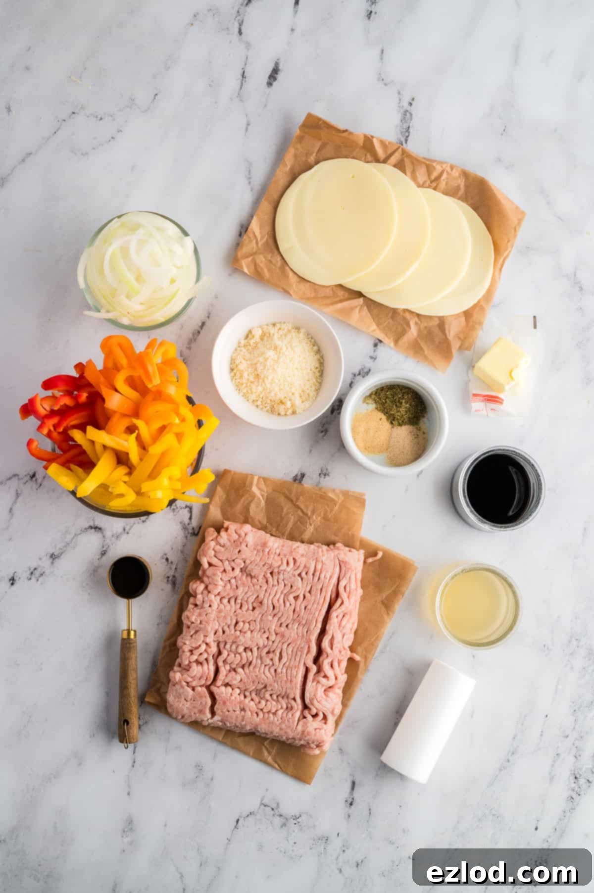Individual ingredients for chicken cheesesteak skillet laid out on a marble background, including ground chicken, peppers, onions, and cheeses.