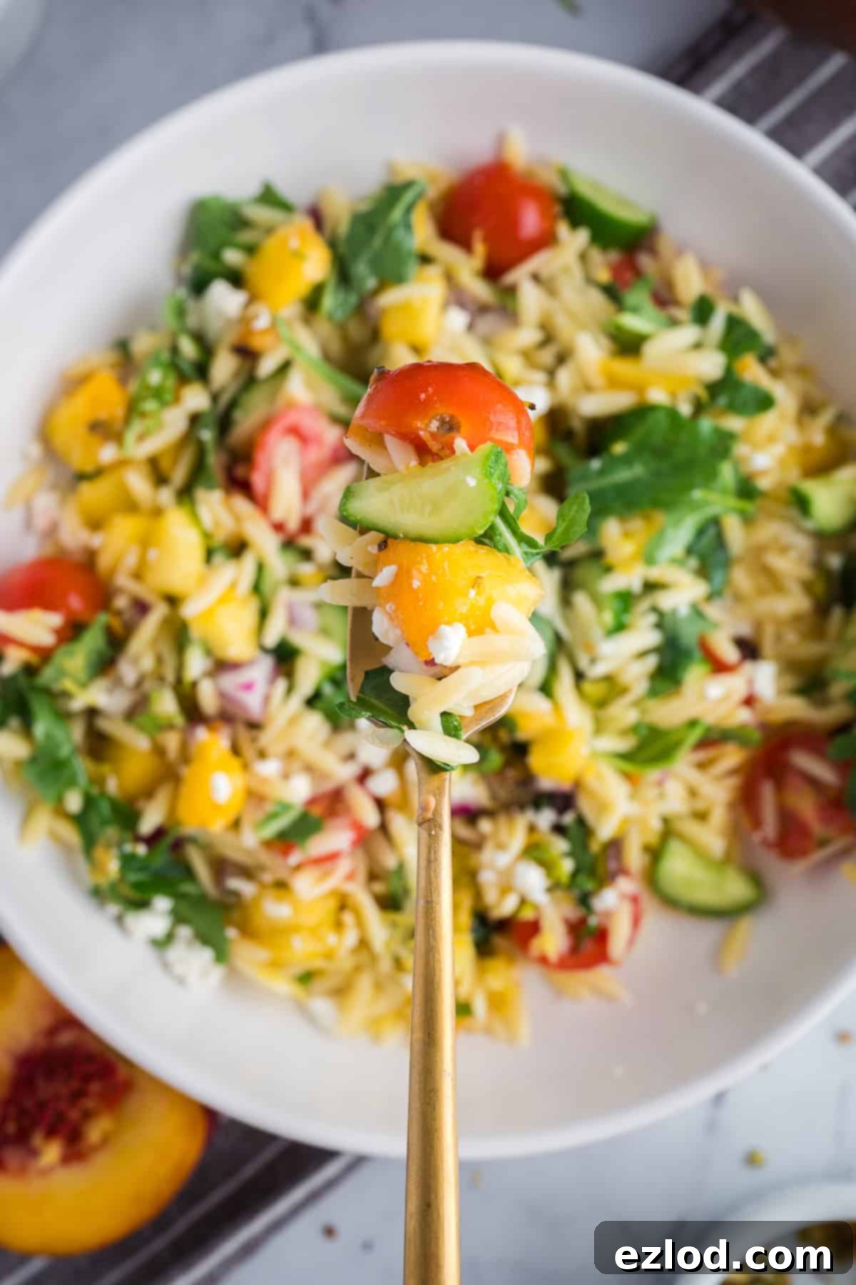 Close-up of a spoon of orzo salad highlighting juicy peaches, creamy feta, cherry tomatoes, cucumbers, and fresh mint.