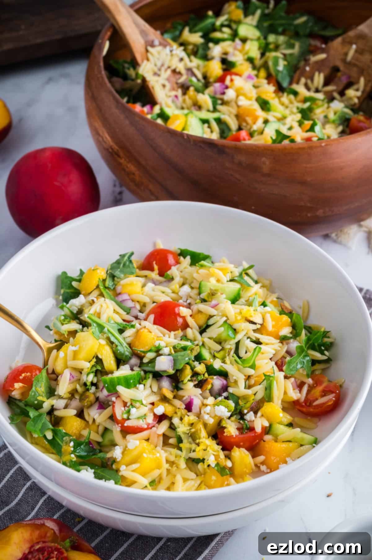 Individual serving of summer orzo salad in a white bowl beside a large wooden bowl filled with the remaining salad.
