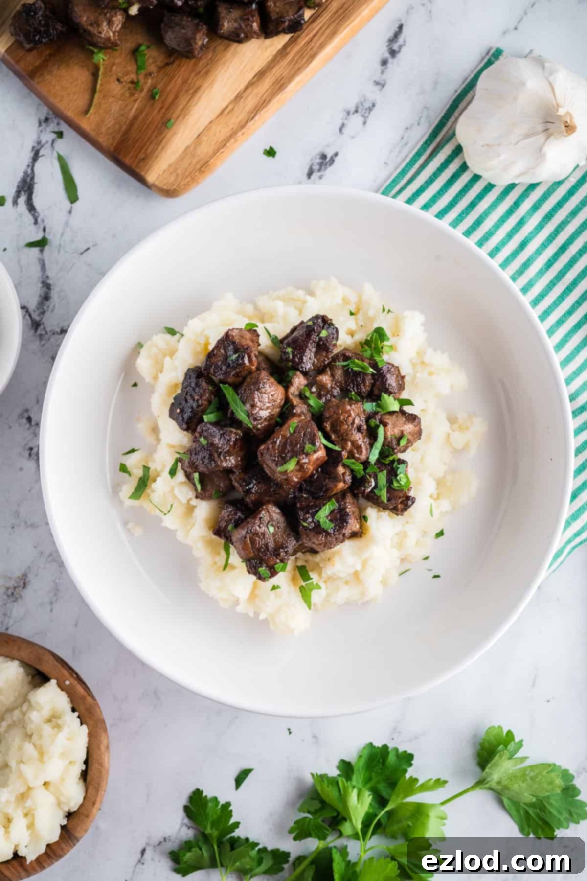 Steak bites over mashed potatoes on a white marble table flanked with a striped kitchen towel.