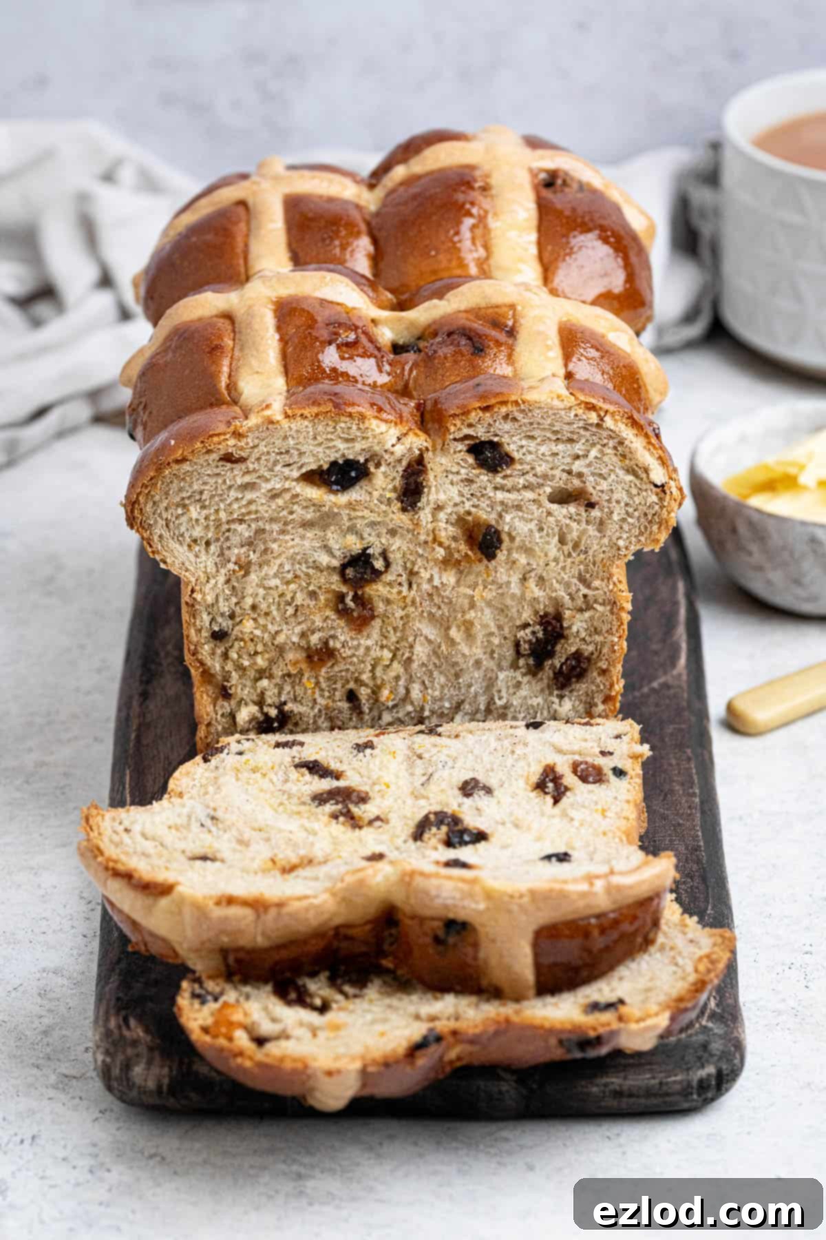 Sliced vegan hot cross bun loaf on a wooden board with a cup of tea and a bowl of butter.