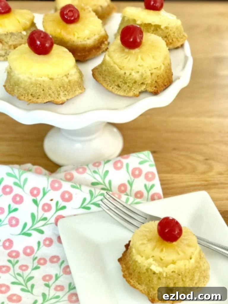 Cupcakes topped with pineapples and cherries on a white cake stand with one cupcake on a square plate with a fork.