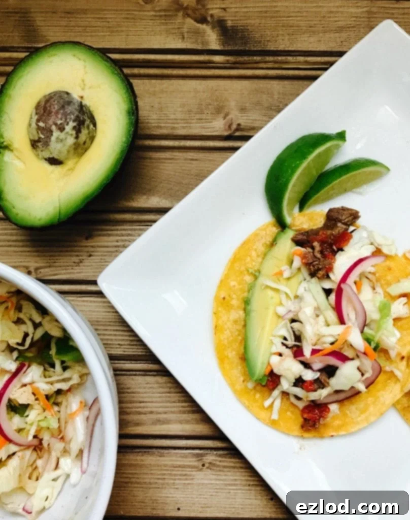 Tender Instant Pot Flank Steak Tacos 5 An overhead view of a single Instant Pot Flank Steak Taco on a white plate, surrounded by fresh lime wedges and a side bowl of cilantro lime coleslaw. The taco is generously filled with tender steak, slaw, avocado, and cilantro, ready to be enjoyed.