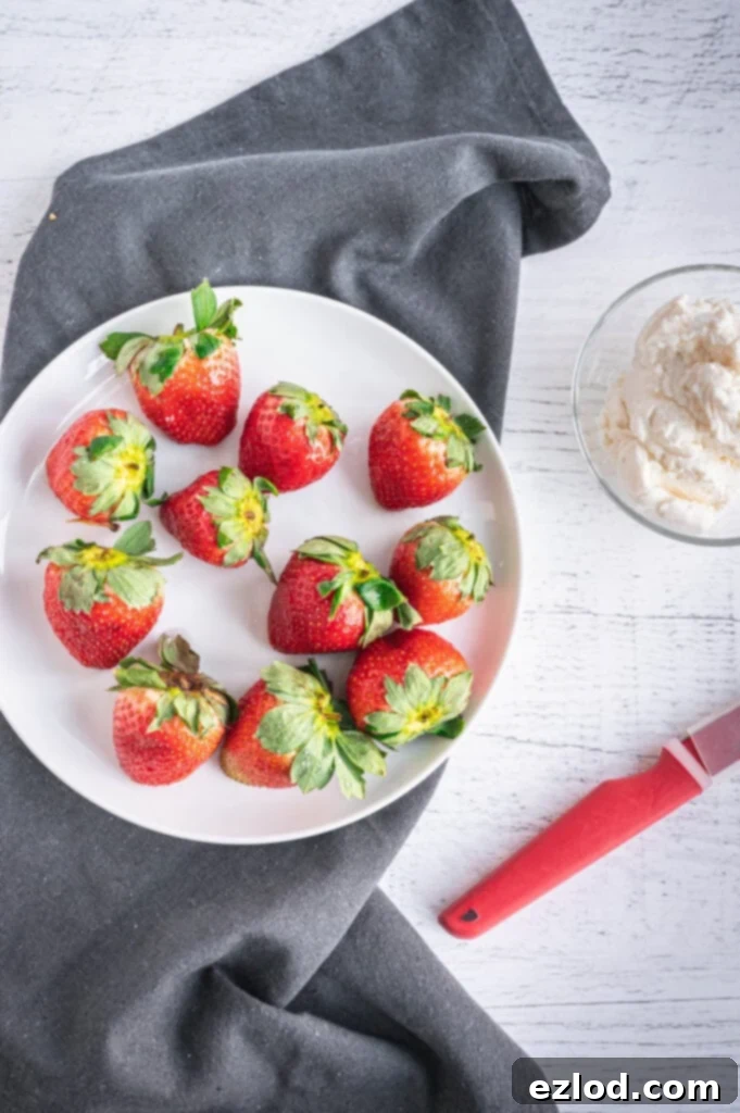 Overhead photo of whole strawberries on a white plate, a red paring knife, and a small glass dish of sweet whipped ricotta on a wooden background with a gray dish towel under the plate.