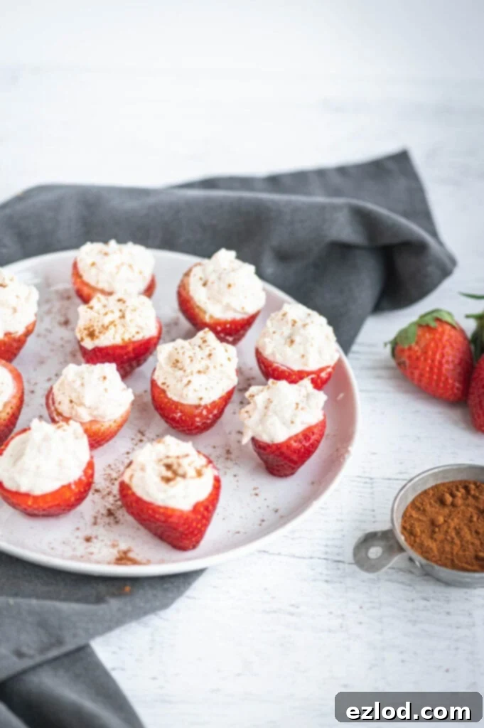 High angle photo of cheesecake stuffed strawberries standing on end on a white plate. There is a bowl of cinnamon and whole strawberries off to the side with a gray towel.