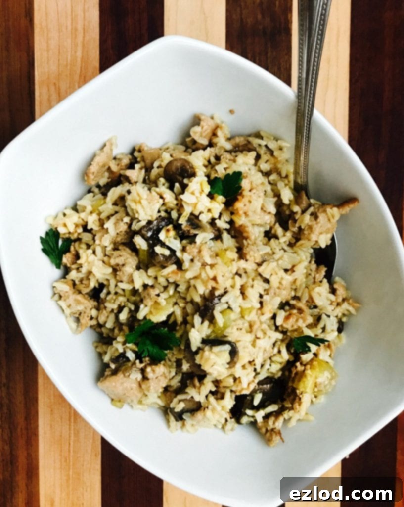 An overhead photo of a white rectangular bowl full of gluten-free Instant Pot Brown Rice and Sausage Stuffing, packed with colorful veggies like celery and mushrooms and topped with fresh parsley, on a striped wooden background.