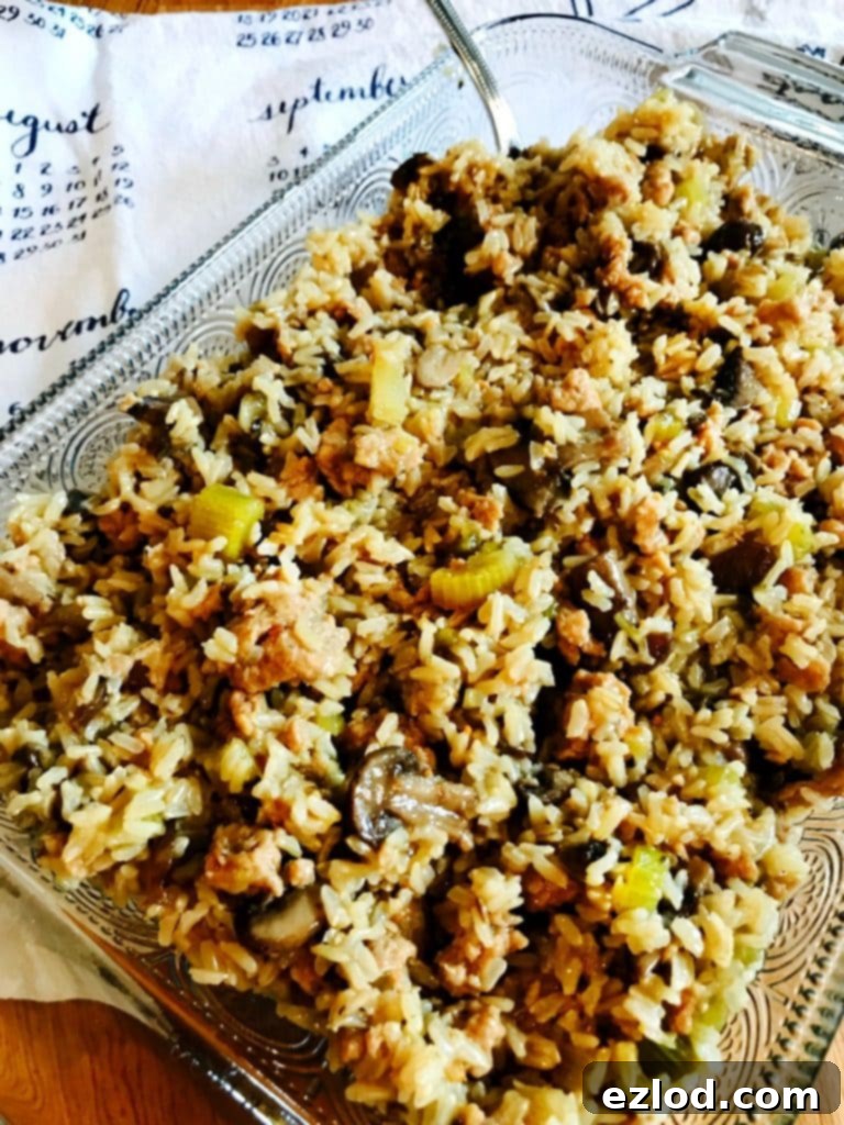 Rectangular glass baking dish full of Instant Pot Brown Rice Stuffing with a silver serving spoon on the side, ready to be enjoyed. In the background is a white and blue tea towel depicting a calendar.