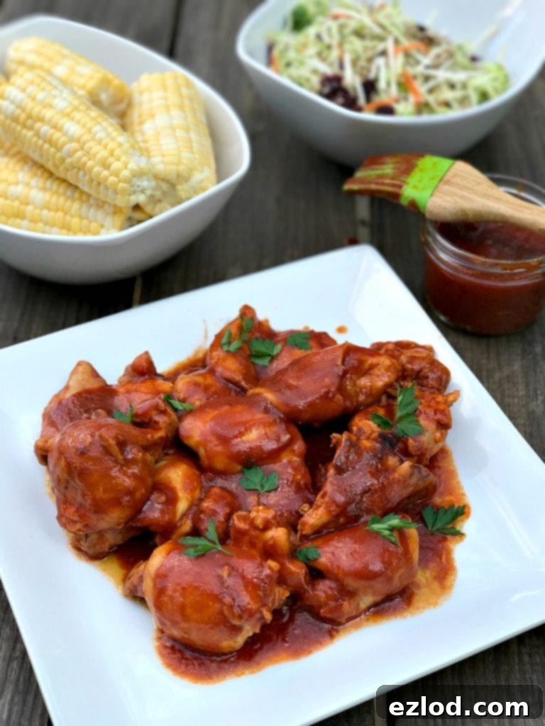 Maple chicken thighs in BBQ sauce on a square white plate. BBQ sauce, corn on the cob, and broccoli slaw in dishes in the background.