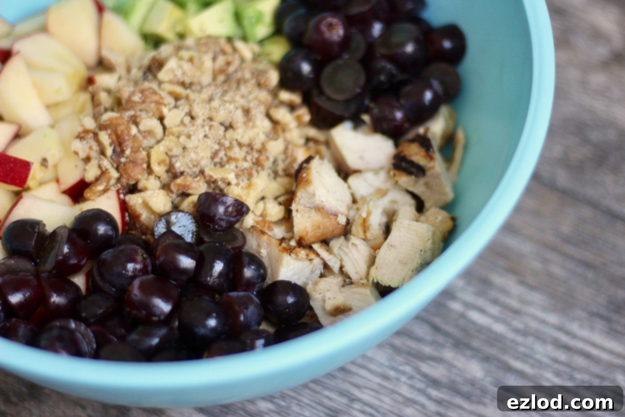 Diced chicken, sliced grapes, diced walnuts, diced apples, and diced avocados sit unmixed in a blue bowl on a wooden surface, ready to be combined into a healthy chicken salad.