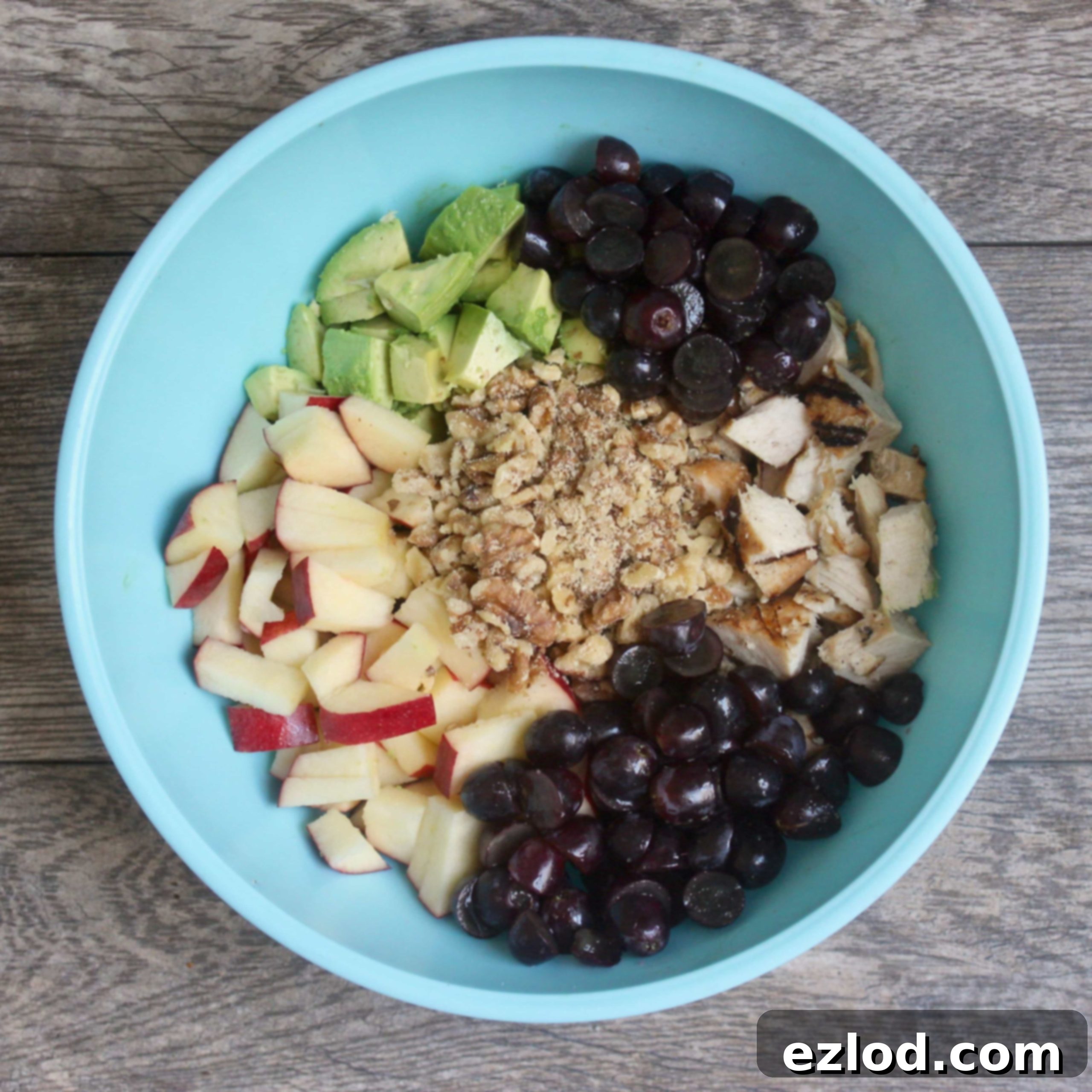 Ingredients for avocado chicken salad (diced chicken, grapes, walnuts, apples, avocado) arranged in a blue bowl before mixing.